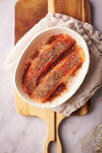 Two raw salmon fillets coated in olive oil and blackening seasoning in a shallow oval bowl.