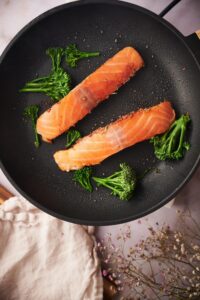 Two salmon fillets cooking spice-side down in a black pan. Pieces of broccolini surround the salmon, cooking in the same pan.