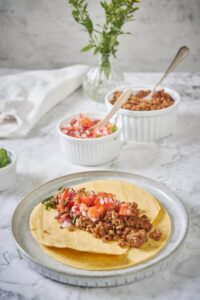 A plate with two fried tortillas stacked one on top of the other. In the center of the top tortilla is a generous serving of chorizo topped with fresh salsa and cilantro. In the background is a bowl of chorizo with a spoon and a smaller bowl of salsa with a small wooden spoon.