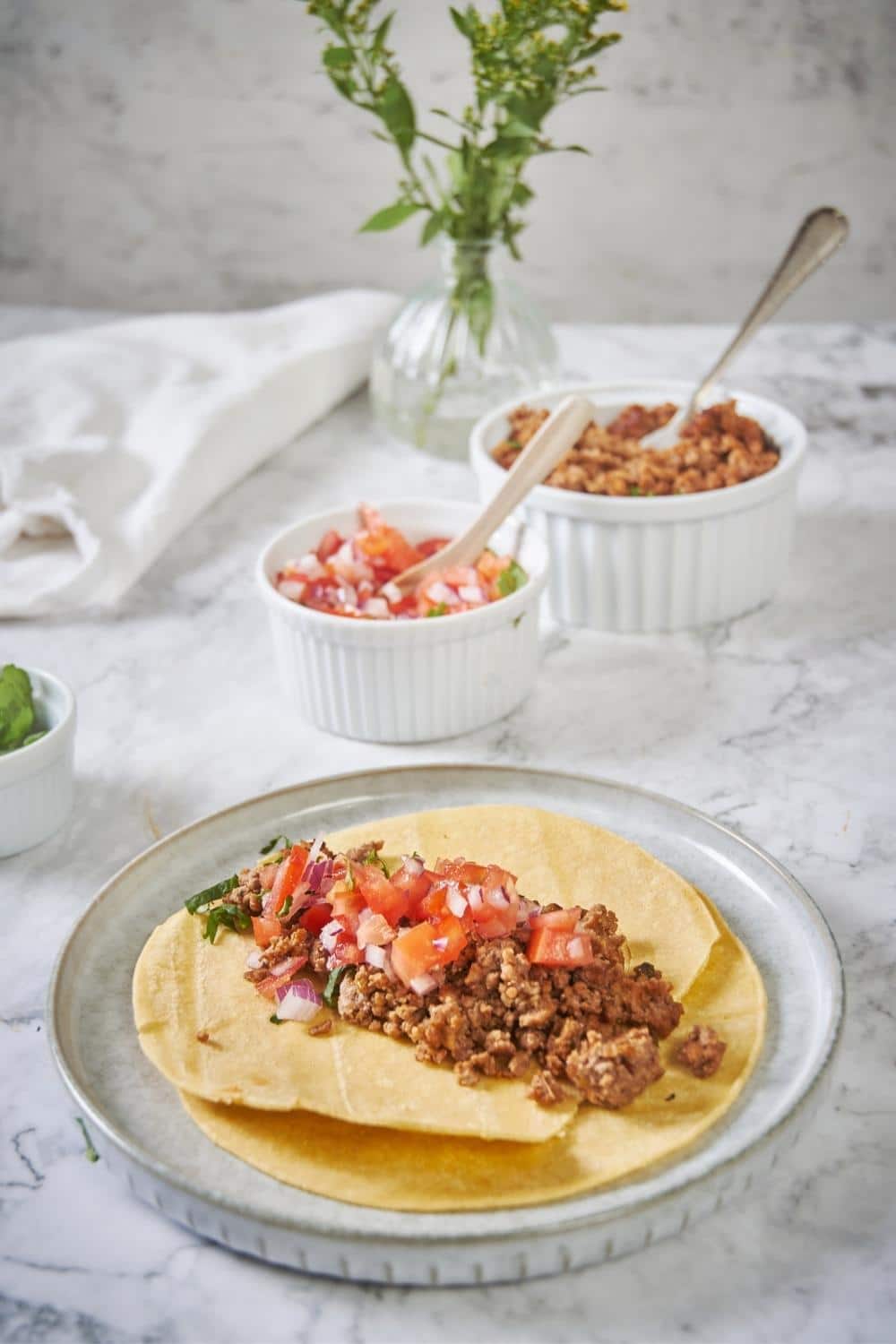 A plate with two fried tortillas stacked one on top of the other. In the center of the top tortilla is a generous serving of chorizo topped with fresh salsa and cilantro. In the background is a bowl of chorizo with a spoon and a smaller bowl of salsa with a small wooden spoon.