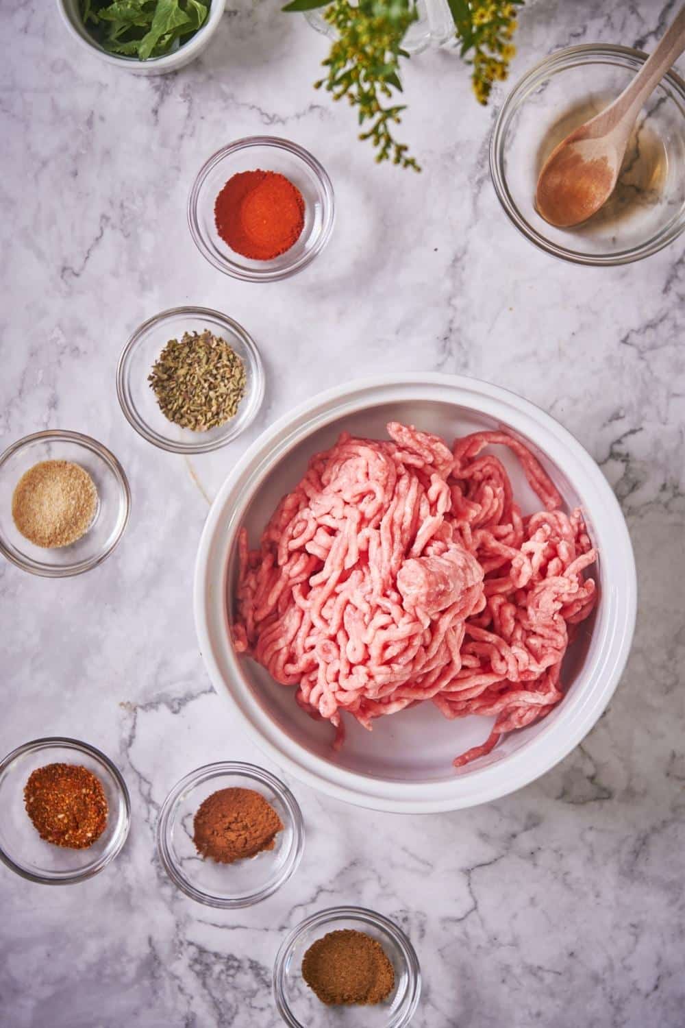 A large bowl of raw ground pork surrounded by smaller glass bowls with individual spices and a glass bowl of red wine vinegar with a wooden spoon.