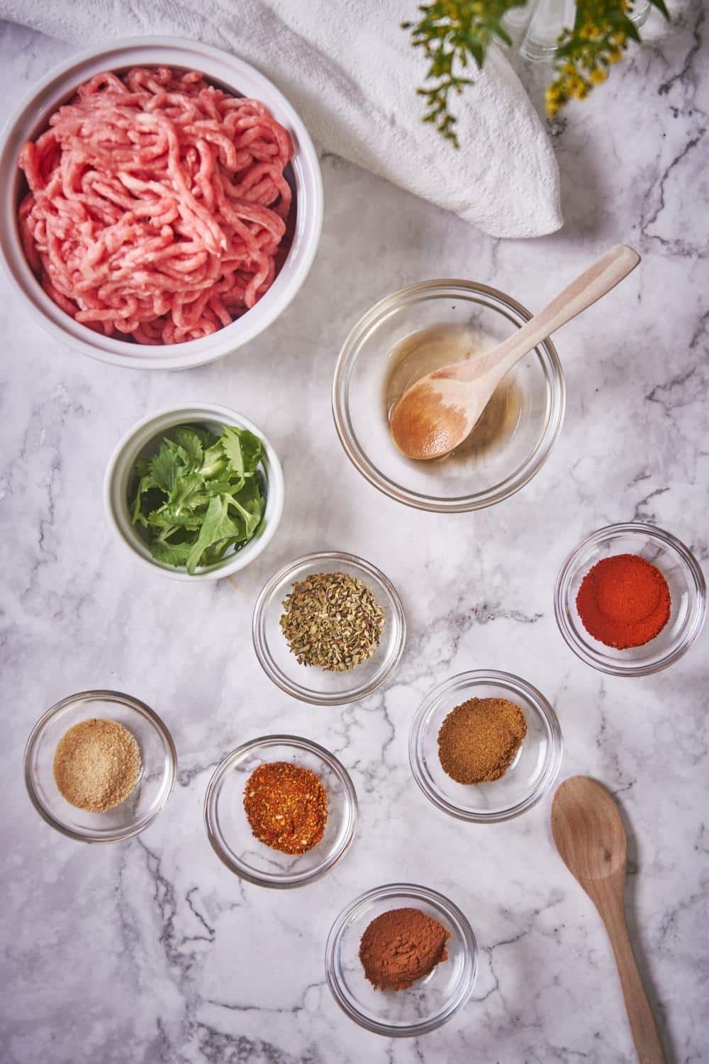 A large bowl of raw ground pork, a small ramekin of chopped cilantro, and small glass bowls each with their own spices: paprika, cumin, garlic powder, cinnamon, Mexican seasoning mix, and red wine vinegar.