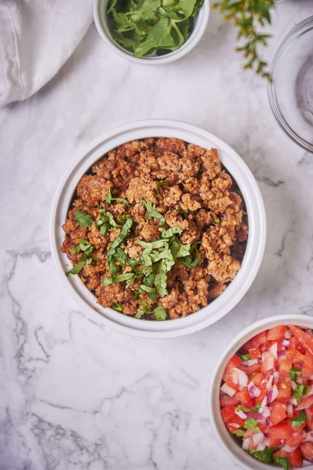 Top shot of a bowl of cooked chorizo next to a bowl of fresh salsa and a bowl of cilantro. The chorizo is garnished with cilantro.