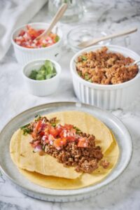 Two fried tortillas are stacked on a plate. The top tortilla has a generous serving of chorizo with fresh salsa and cilantro. In the background is a bowl of cooked chorizo, a bowl of fresh salsa, and a small bowl of chopped cilantro.