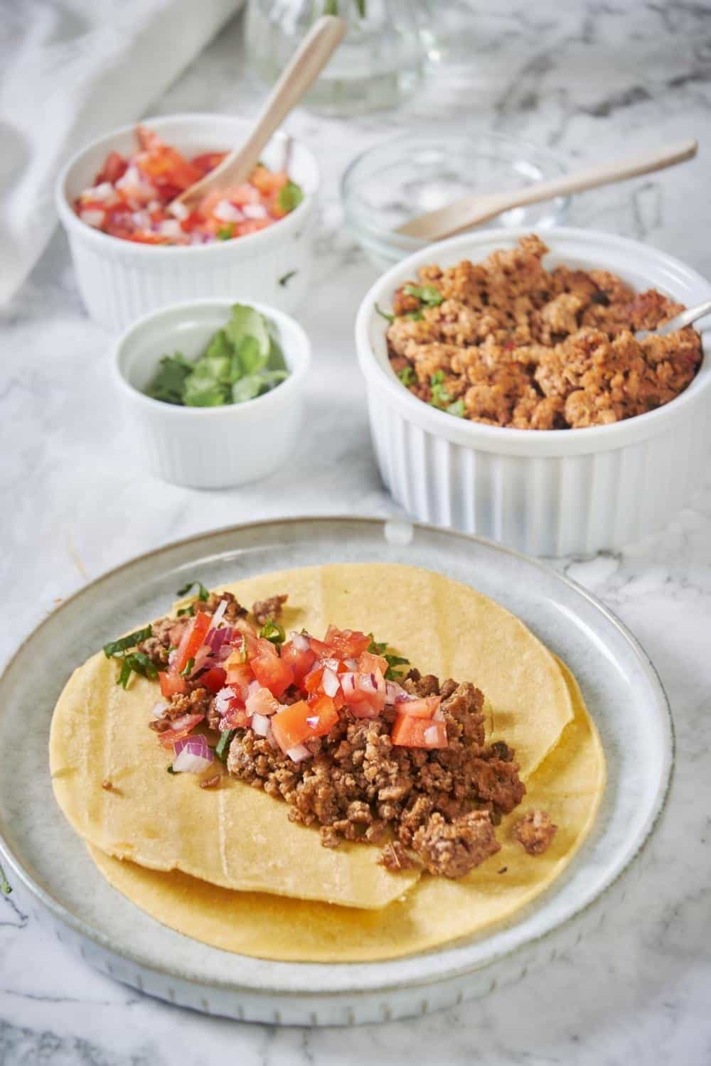 Two fried tortillas are stacked on a plate. The top tortilla has a generous serving of chorizo with fresh salsa and cilantro. In the background is a bowl of cooked chorizo, a bowl of fresh salsa, and a small bowl of chopped cilantro.
