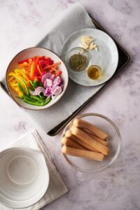 Sliced bell peppers and onions in a white bowl on top of a parchment paper lined baking tray. Next to it is a grey plate with sliced garlic and smaller bowls of herbs and olive oil. Outside the tray is a glass bowl of uncooked chicken sausages.