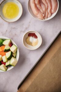 A small bowl of spices surrounded by larger bowls of chopped veggies, raw chicken tenderloins, melted butter, and a parchment paper lined baking sheet.