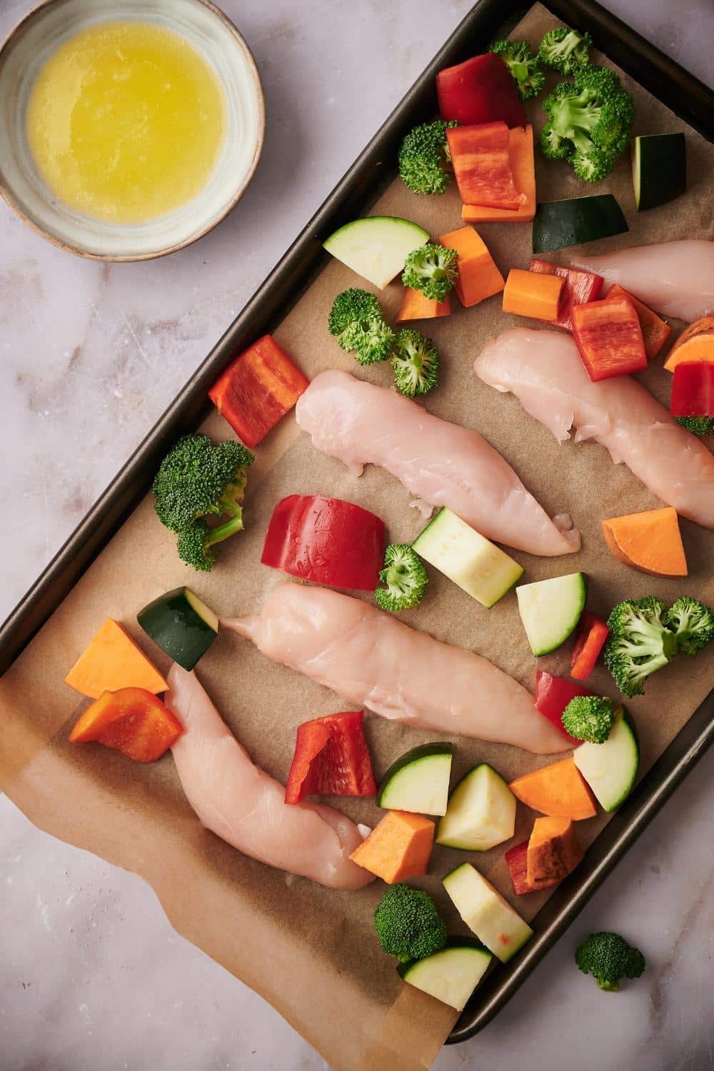 5 raw chicken tenderloins surrounded with chopped veggies in a parchment paper lined sheet tray. A small bowl of melted butter is on the side.