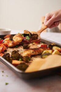 A wooden spatula scooping a baked chicken tenderloin with veggies from a parchment paper lined sheet pan.