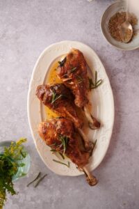 Three roasted turkey legs on an oval plate, garnished with rosemary leaves and golden cooking juices. A small bowl of spice mix with a metal spoon is on the side.