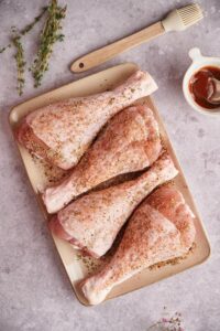 raw turkey legs coated in seasoning mix on a rectangle plate. A silicone pastry brush can be seen on the side as well as a small bowl of BBQ sauce with a spoon in it.