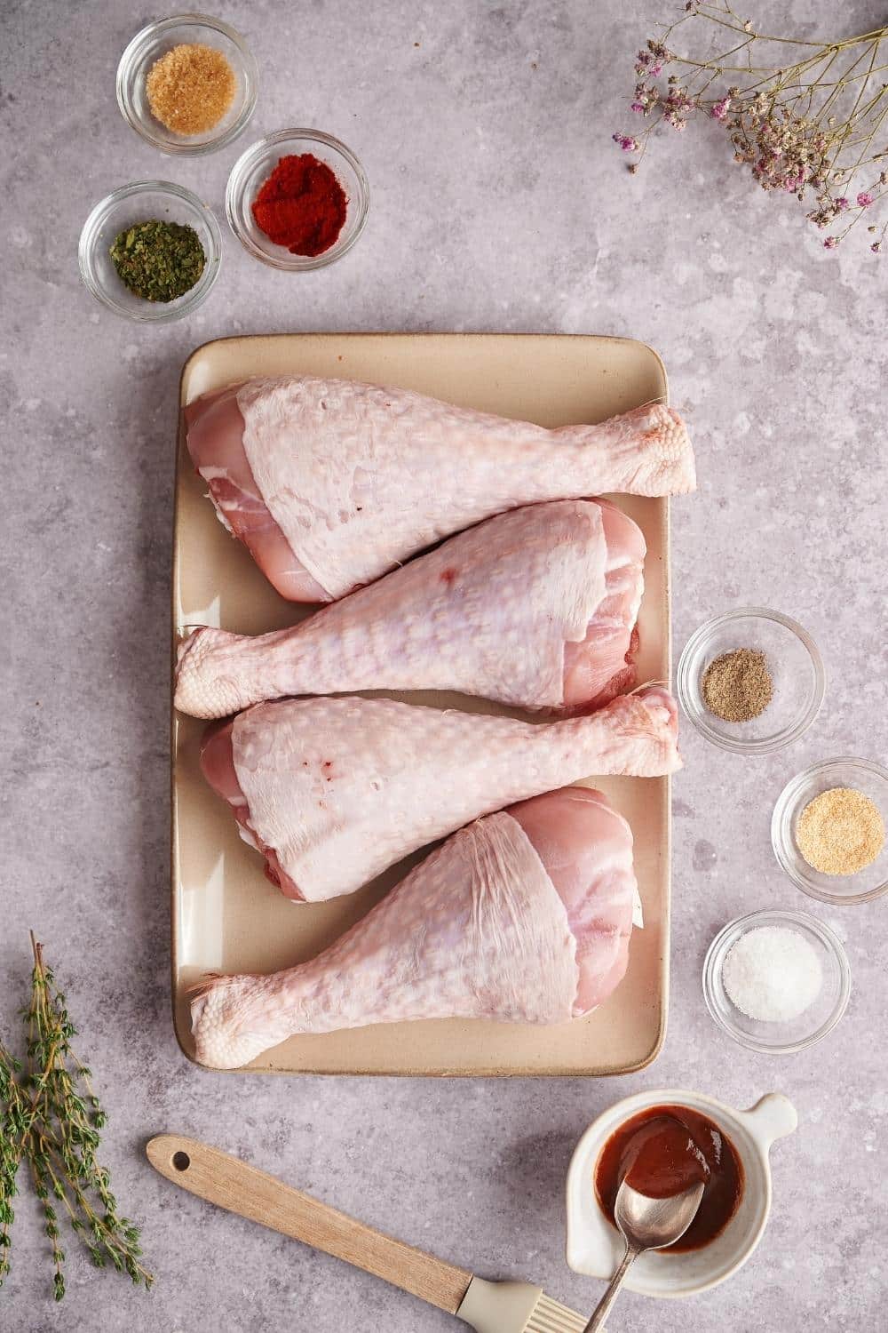 4 raw turkey legs on a rectangular plate surrounded by small glass bowls of garlic powder, smoked paprika, dried oregano, onion powder, salt, black pepper, and BBQ sauce, as well as a pastry brush and some fresh thyme.