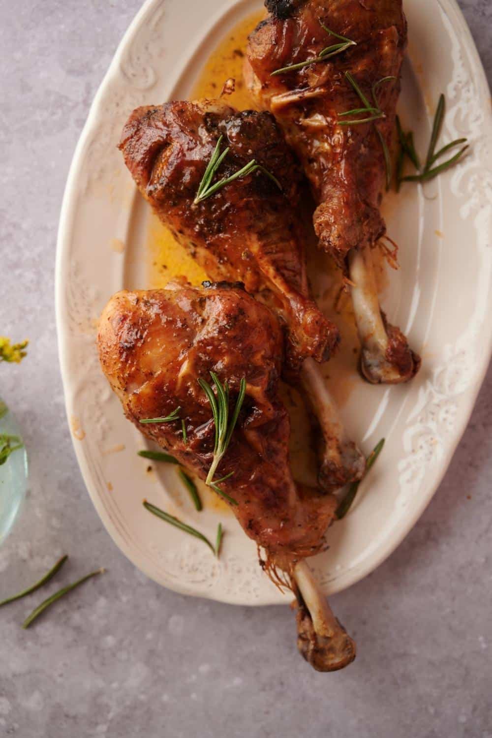 Three roasted turkey legs on an oval cream-colored plate. The turkey legs are coated in cooking juices and garnished with fresh rosemary leaves.