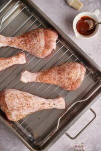 Four seasoned raw turkey legs on a roasting tray. A small bowl of BBQ sauce with a metal spoon can be seen on the side next to part of a pastry brush.