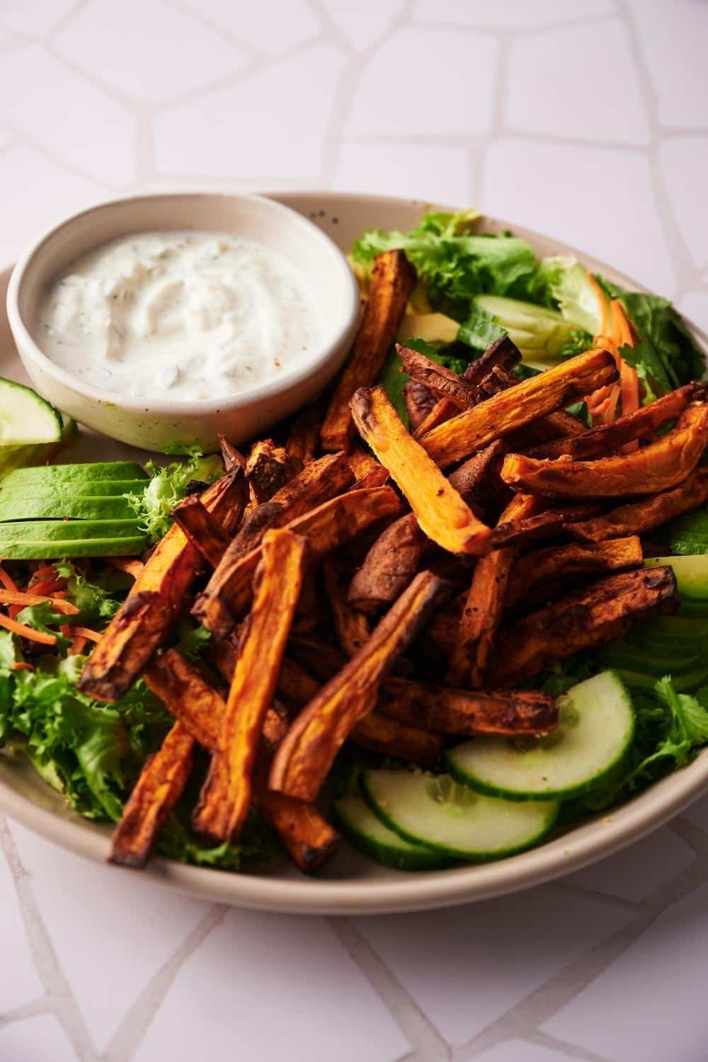 Air fried sweet potato fries on a beige plate. The fries are resting on top of a salad of lettuce and sliced cucumbers next to a small bowl of creamy dipping sauce.