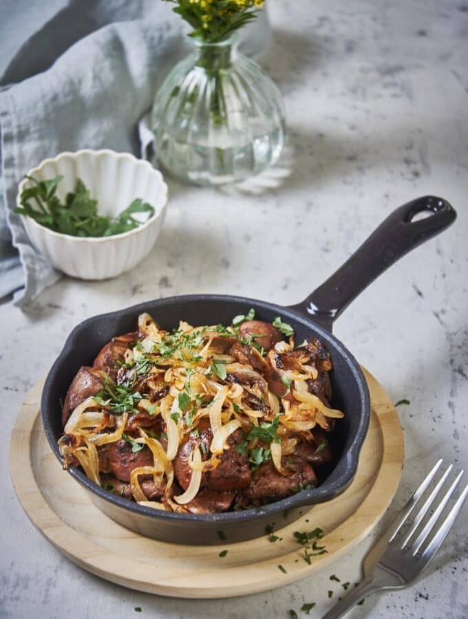 sauteed chicken livers and onions garnished with parsley in a small cast iron pan set on a wooden plate. A fork sits next to the pan and a tea towel, vase of flowers, and small bowl of parsley can be seen in the background.