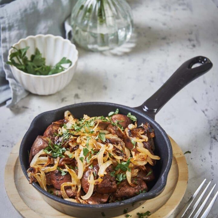 sauteed chicken livers and onions garnished with parsley in a small cast iron pan set on a wooden plate. A fork sits next to the pan and a tea towel, vase of flowers, and small bowl of parsley can be seen in the background.