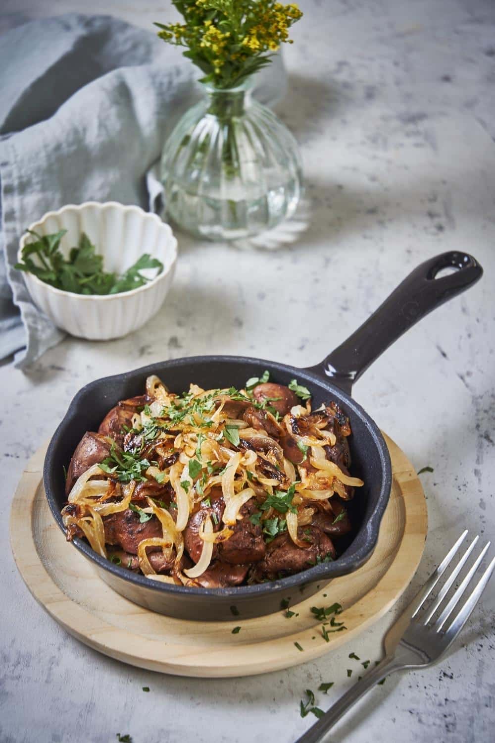 sauteed chicken livers and onions garnished with parsley in a small cast iron pan set on a wooden plate. A fork sits next to the pan and a tea towel, vase of flowers, and small bowl of parsley can be seen in the background.