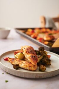 Baked chicken tenderloins on a bed of roasted veggies arranged on a plate. A parchment paper lined sheet pan with more chicken tenders and veggies can be seen at the back next to a small bowl of herbs.