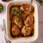 Overhead view of a baking dish filled with 6 baked chicken thighs garnished with parsley and sitting in golden cooking juices. A spoon rests on top, ready to take out one of the chicken thighs.