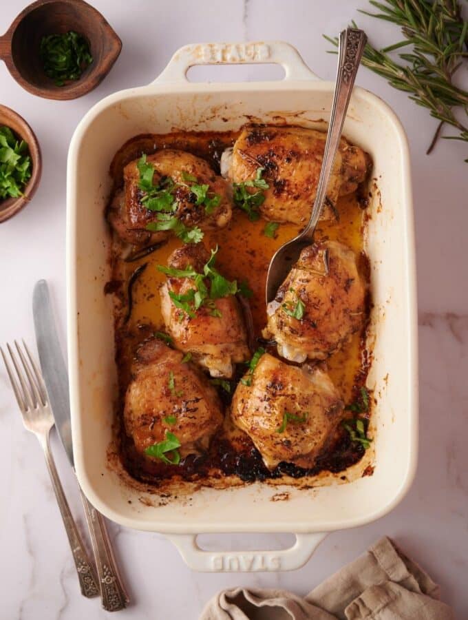 Overhead view of a baking dish filled with 6 baked chicken thighs garnished with parsley and sitting in golden cooking juices. A spoon rests on top, ready to take out one of the chicken thighs.