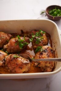a baking dish with 6 baked chicken thighs garnished with herbs and sitting in golden cooking juices. A spoon is taking out one of the chicken thighs