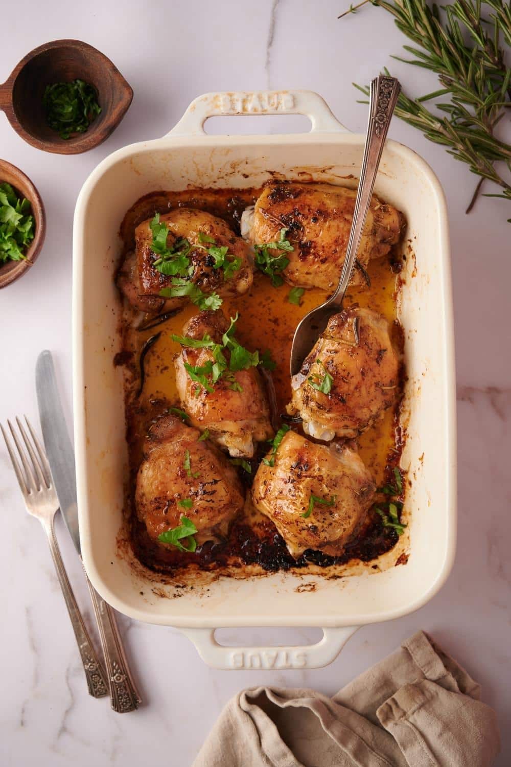 Overhead view of a baking dish filled with 6 baked chicken thighs garnished with parsley and sitting in golden cooking juices. A spoon rests on top, ready to take out one of the chicken thighs.