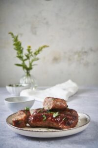 Roasted brats on a white plate. One of the brats is cut into three pieces and arranged on top of three roasted brats. Next to the plate is mustard in a small bowl.