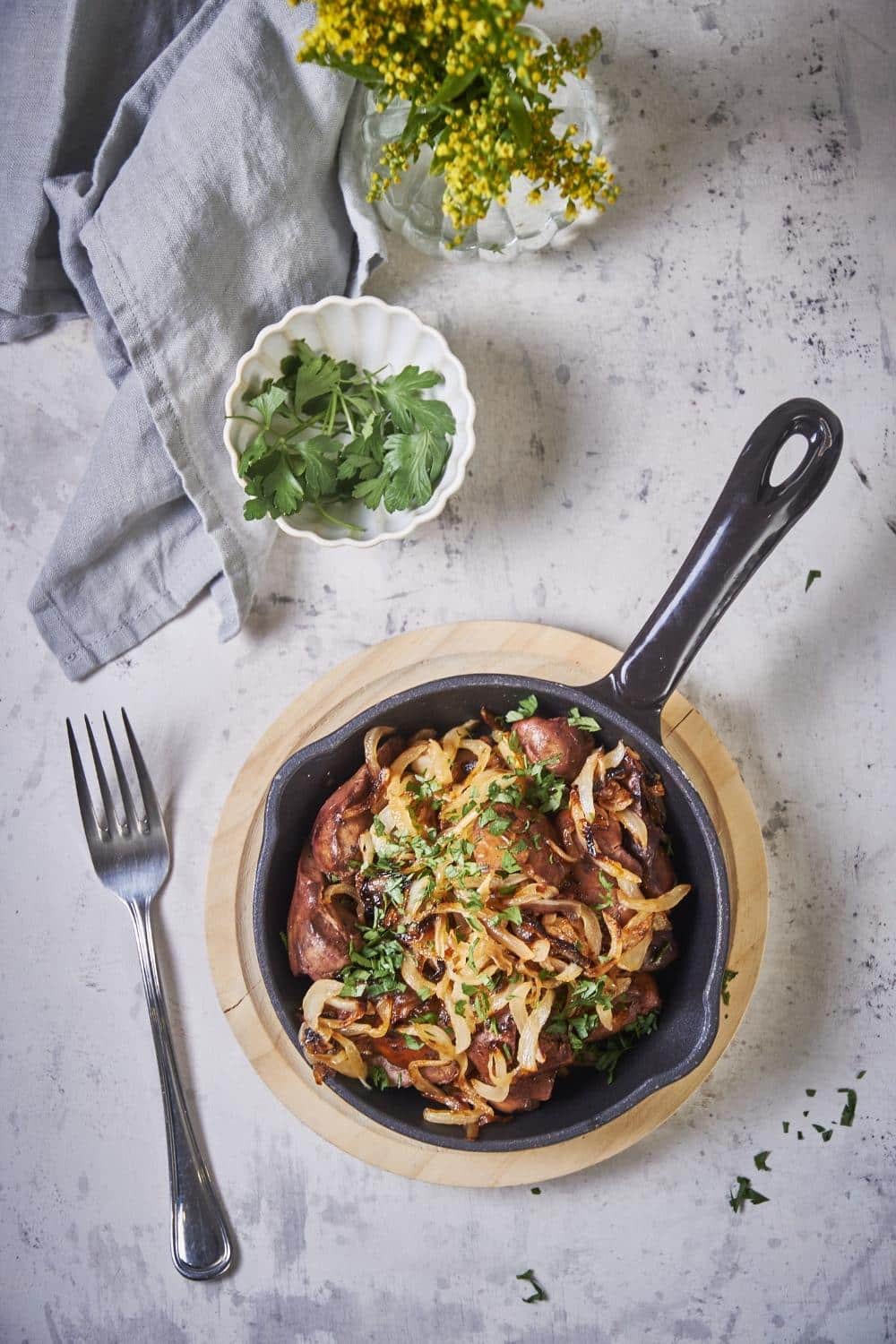 Top view of sauteed chicken livers and onions in a small cast iron pan on a wooden plate. A fork sits next to the pan and a bowl of parsley for garnish is on the side.