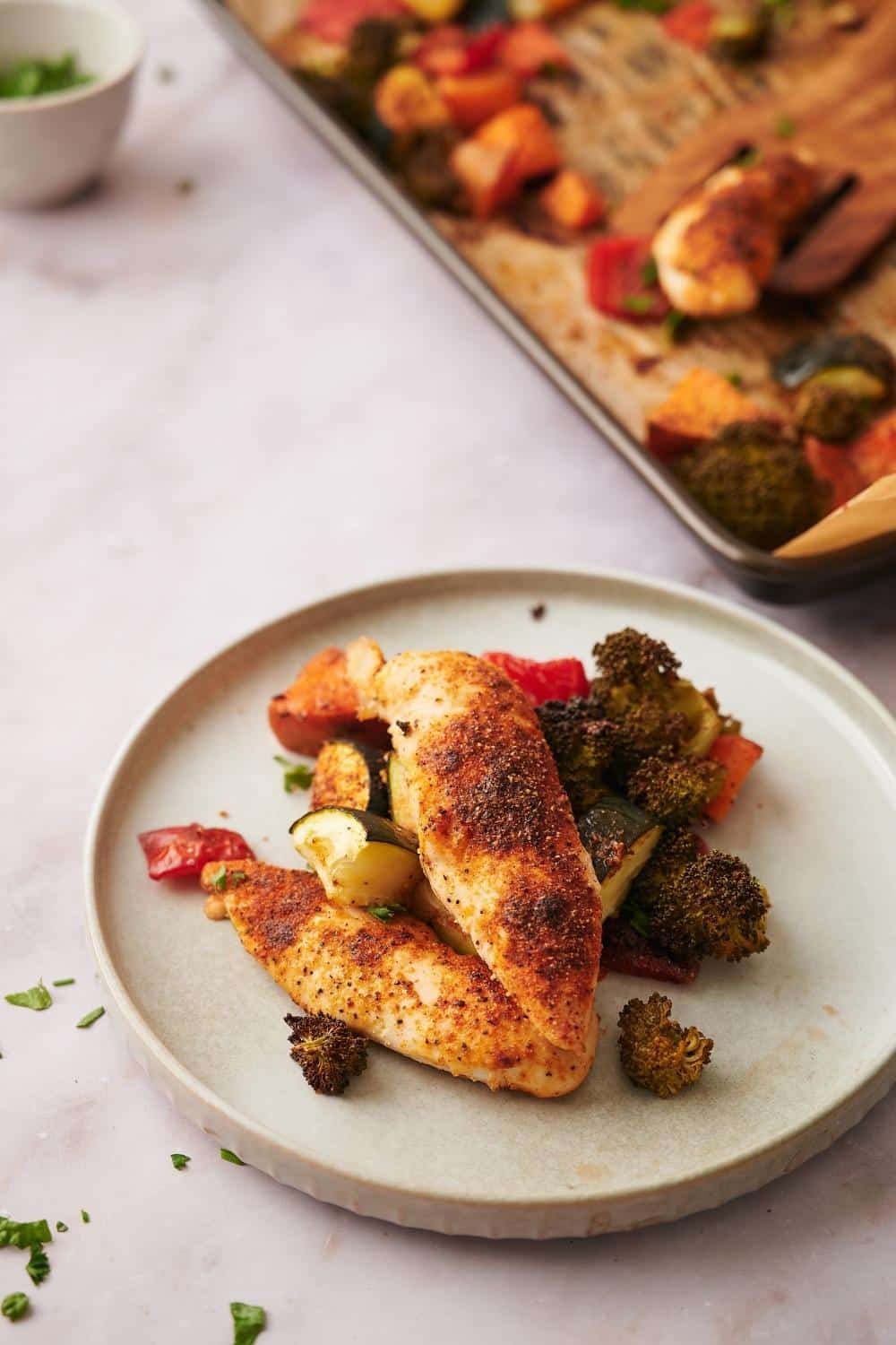 Baked chicken tenderloins on top of roasted chopped veggies on a plate. They're garnished with herbs, some of which are delicately scattered on the table top. Part of a bowl of herbs and a parchment paper lined baking sheet with more chicken and veggies can be seen in the back.