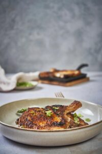 seared thin pork chops in an ecru plate on top of cooked asparagus. In the background is a pan of more seared thin pork chops and a small plate of chopped green onions.