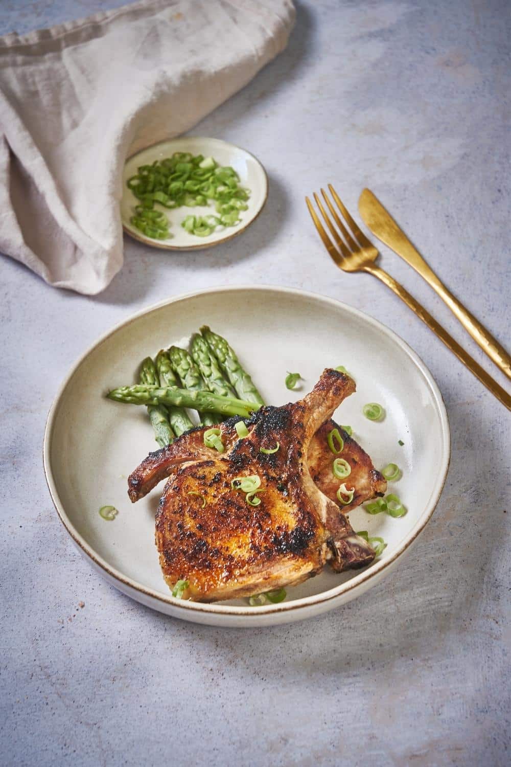 seared thin pork chops garnished with chopped green onion on a bed of cooked asparagus on an ecru plate. Next to the plate is a gold fork and knife and behind is a small saucer of chopped green onions and a tea towel.