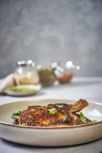 Seared thin pork chops over cooked aspargus in an ecru plate. In the background is three round glass spice jars with garlic powder, pepper, and paprika, and a small plate of chopped green onions.