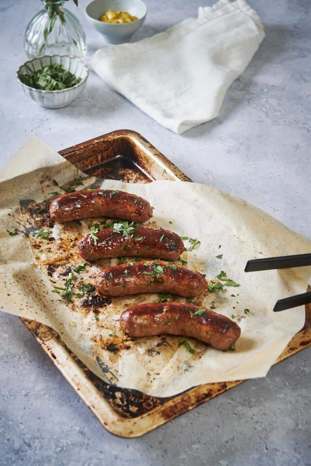 Four roasted brats garnished with parsley on a sheet pan lined with parchment paper. A pair of black tongs is on the corner of the pan.