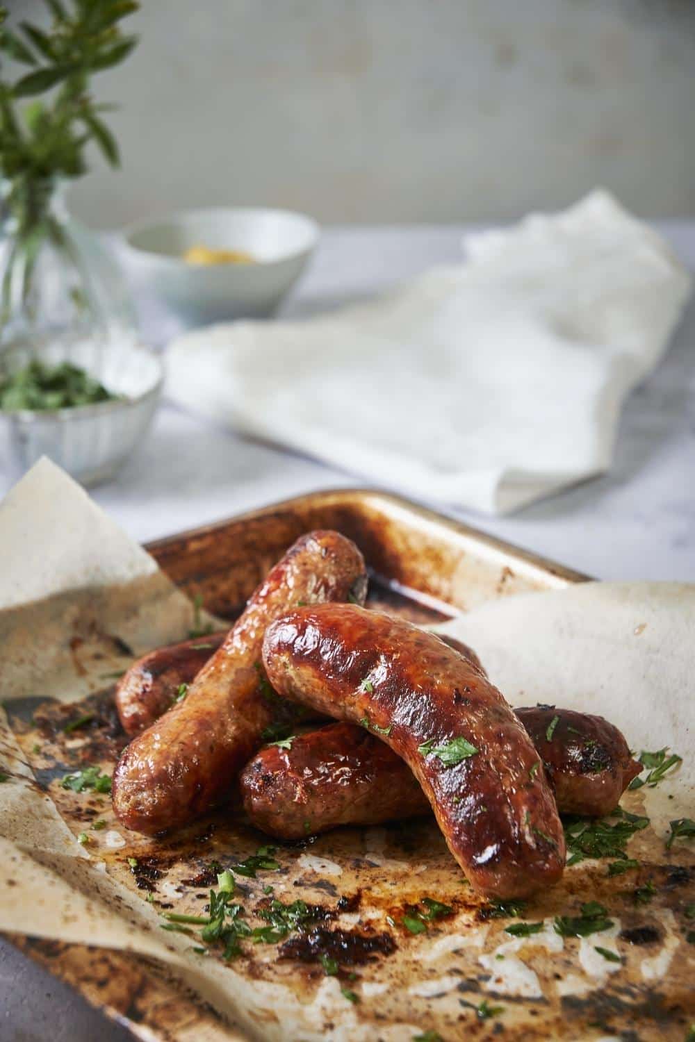 Four roasted brats slightly stacked on a sheet pan lined with parchment paper. The brats are garnished with parsley.