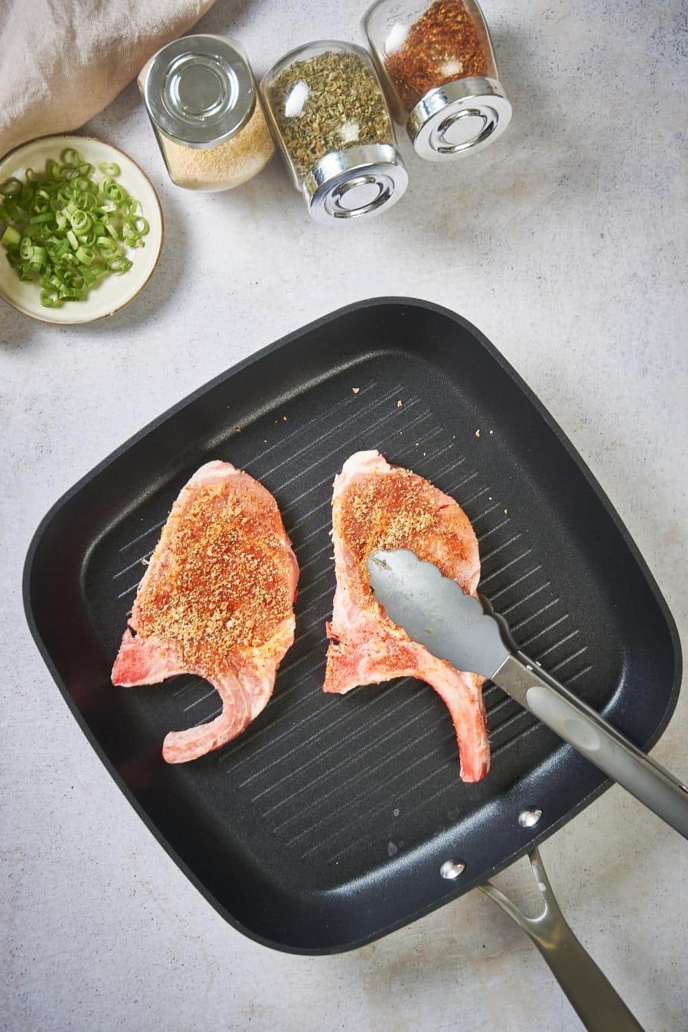 Two seasoned raw pork chops in a grill pan. One pork chop is being placed with metal tongs. Next to the pan are three spice jars of garlic powder, black pepper, and paprika, and a small plate of green onion garnish.