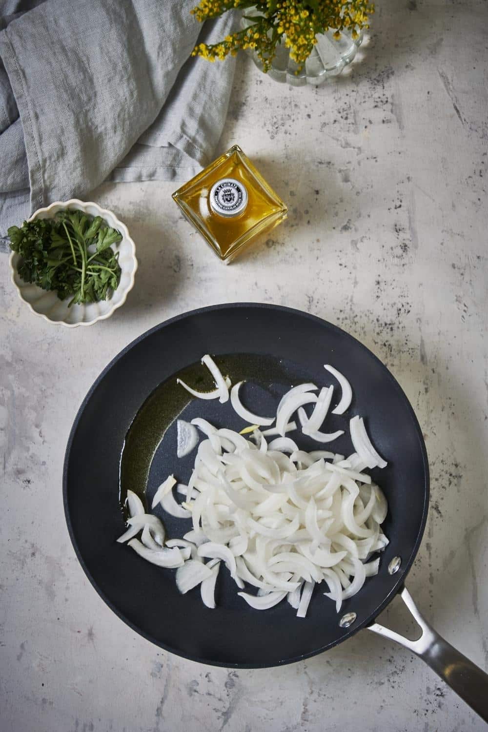 Raw sliced onions in a black skillet with olive oil. A bottle of oil and a small bowl of garnish can be seen on the side.