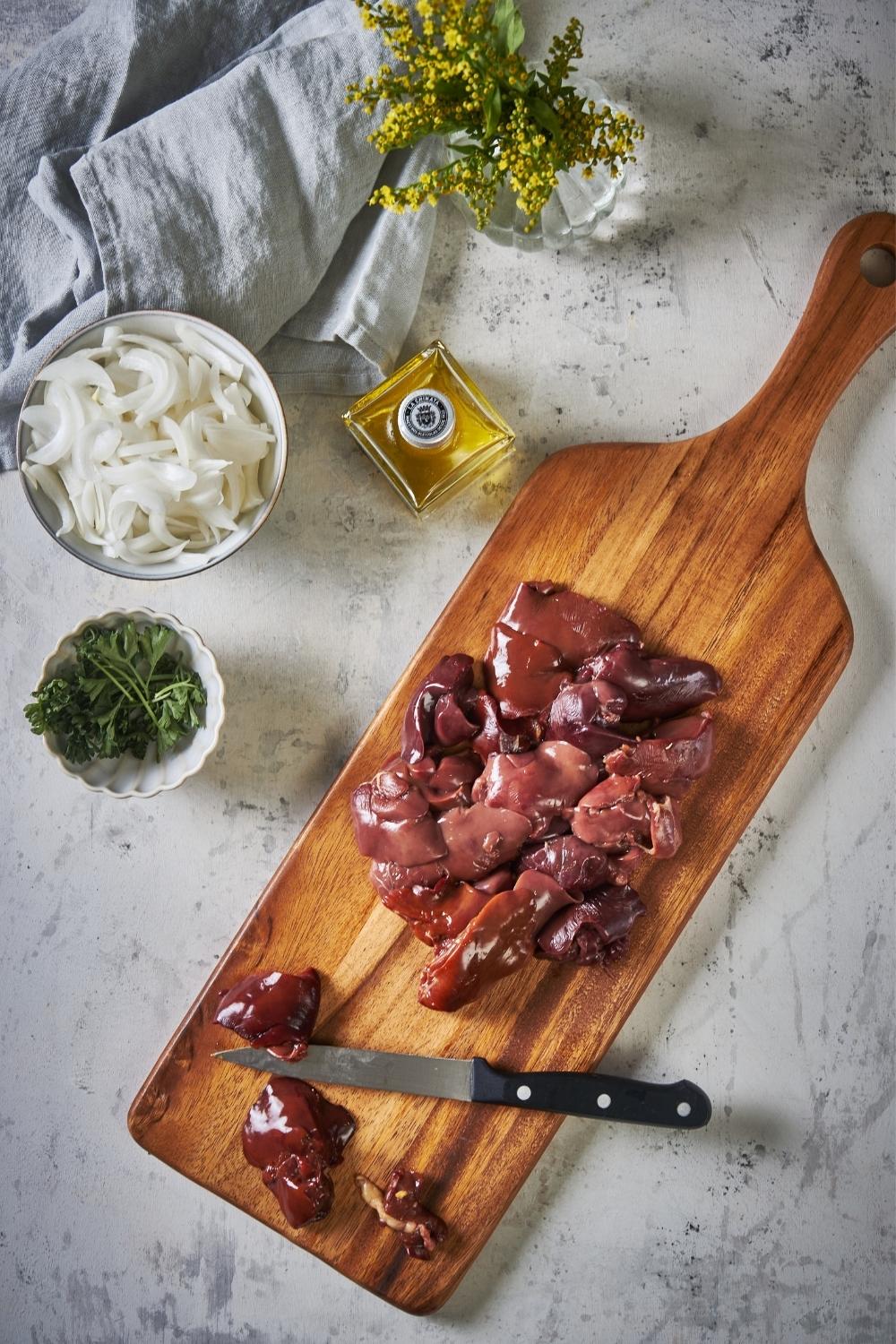 Raw chicken livers on a wooden cutting board. On one side of the board are the sliced chicken livers and on the other side are the remaining whole chicken livers. A small knife is on the cutting board and a bottle of olive oil, bowl of raw sliced onions, and bowl of parsley are to the side.