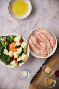 Clockwise from top: A shallow medium bowl of melted butter, 6 raw chicken tenderloins in a large bowl, small glass bowls of garlic powder, paprika, and lemon pepper on a parchment paper lined baking sheet, a small glass bowl of salt, a large bowl of chopped veggies.