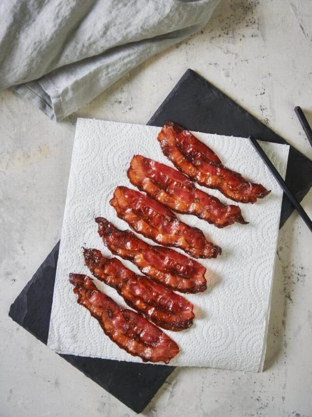 Six pieces of cooked turkey bacon on a paper towel on top of a slate serving platter. A pair of black tong rests on the edge of the platter and a tea towel is folded on the upper left corner.