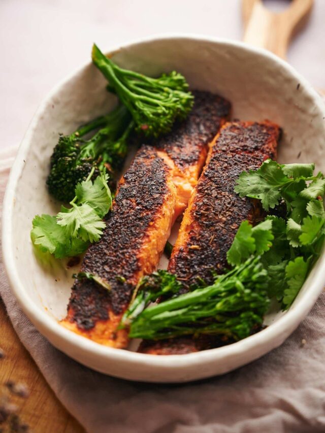 two blackened salmon fillets in a shallow white bowl garnished with seared broccolini and fresh cilantro.