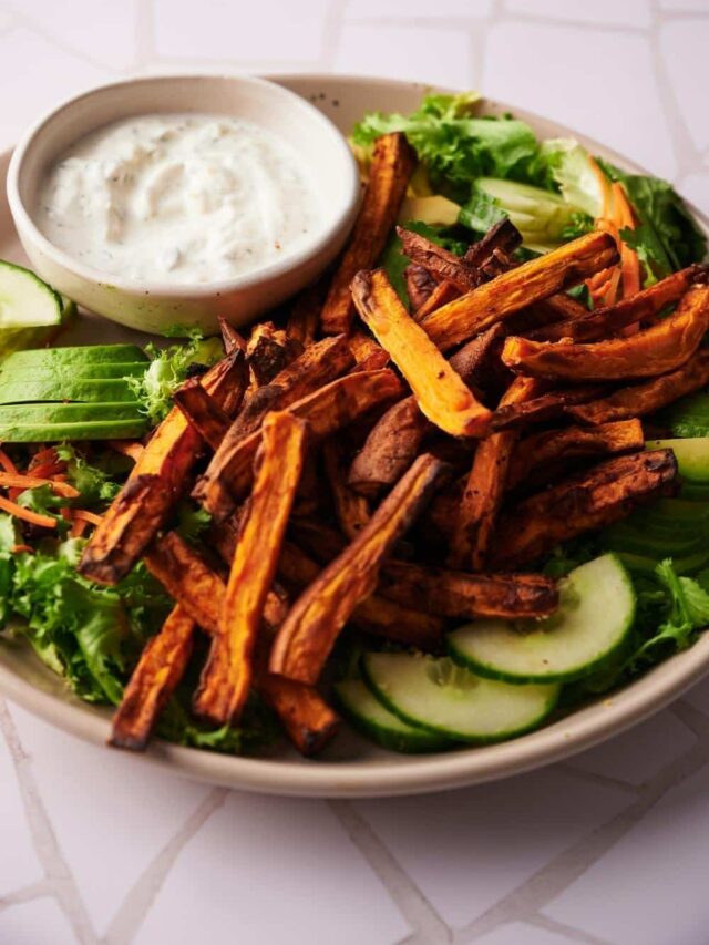 Air fried sweet potato fries on a beige plate. The fries are resting on top of a salad of lettuce and sliced cucumbers next to a small bowl of creamy dipping sauce.