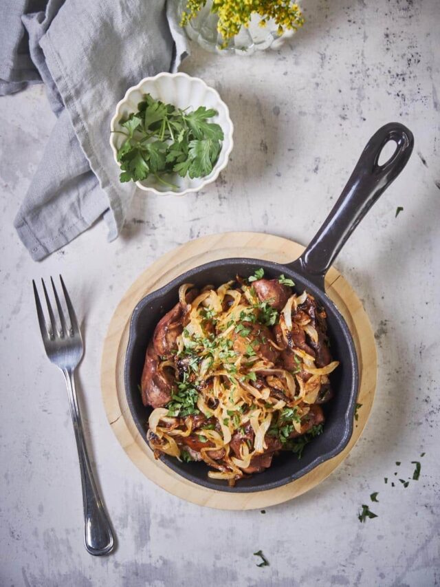 Top view of sauteed chicken livers and onions in a small cast iron pan on a wooden plate. A fork sits next to the pan and a bowl of parsley for garnish is on the side.