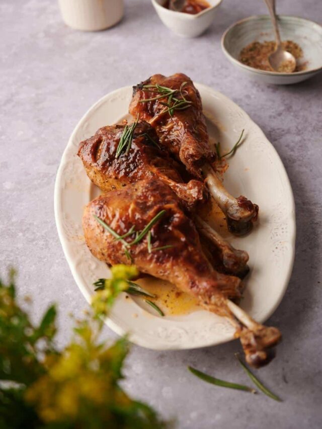 Three roasted turkey legs garnished with cooking juices and rosemary leaves on an oval plate. In the back is a bowl of spice mix and a bowl of BBQ sauce, both with metal spoons in them, as well as a small ceramic pitcher.