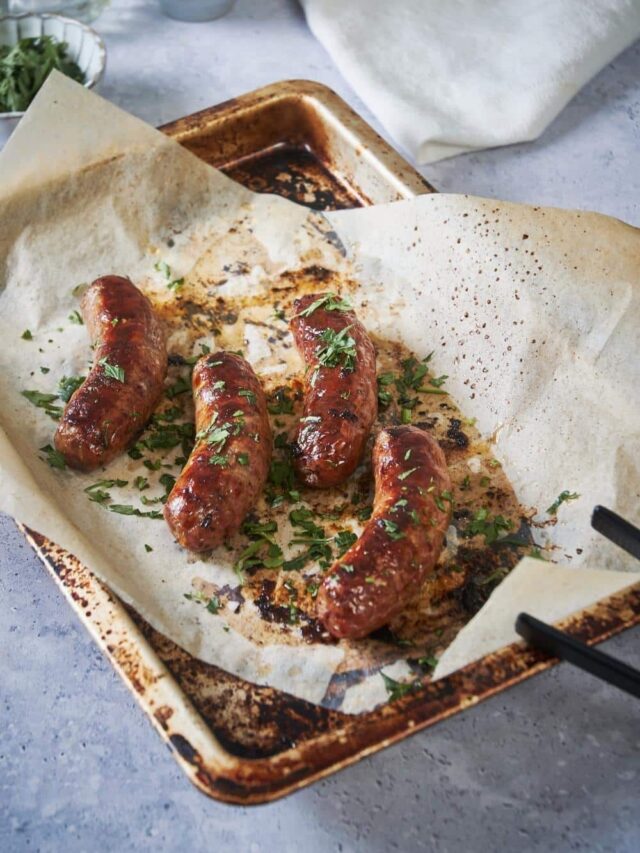 Four roasted brats arranged neatly on a baking sheet lined with parchment paper. The brats are garnished with fresh parsley. A pair of black tongs sits on the corner of the sheet and small bowls of mustard and parsley can be seen in the back.