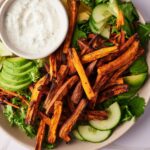 Sweet potato fries on a salad of mixed greens, sliced avocado, and sliced cucumber. A small bowl of creamy dipping sauce is also on the plate.