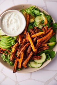 Sweet potato fries on a salad of mixed greens, sliced avocado, and sliced cucumber. A small bowl of creamy dipping sauce is also on the plate.