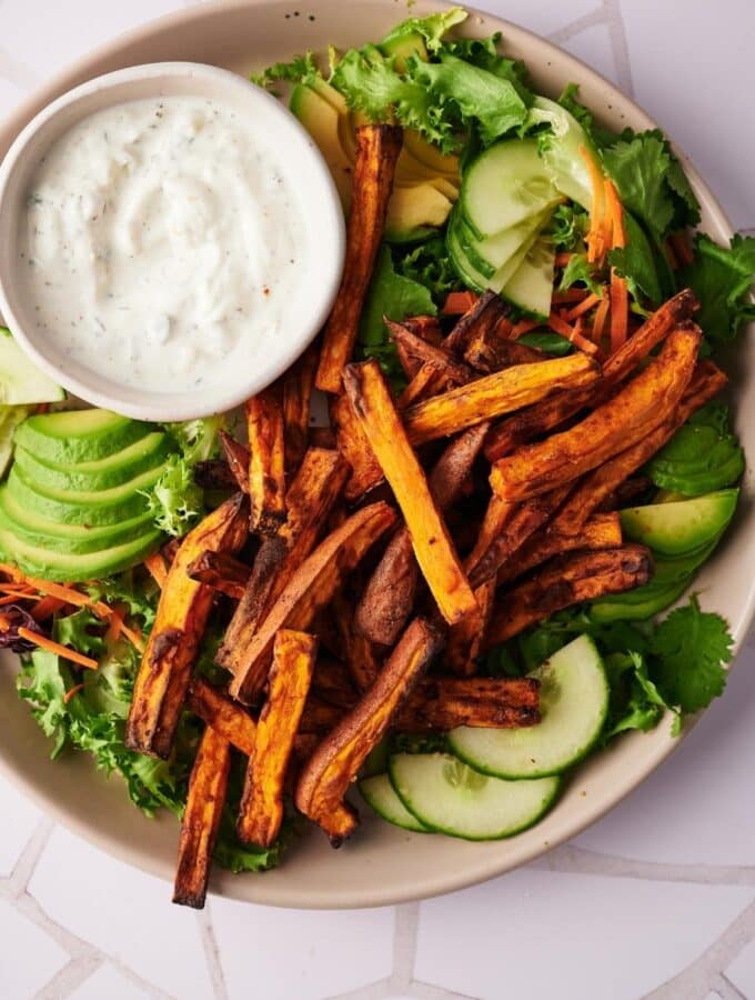 Sweet potato fries on a salad of mixed greens, sliced avocado, and sliced cucumber. A small bowl of creamy dipping sauce is also on the plate.