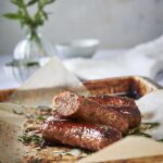 Four brats on a sheet pan lined with parchment paper garnished with parsley. One of the brats is cut in half to show the pink and juicy cross section.