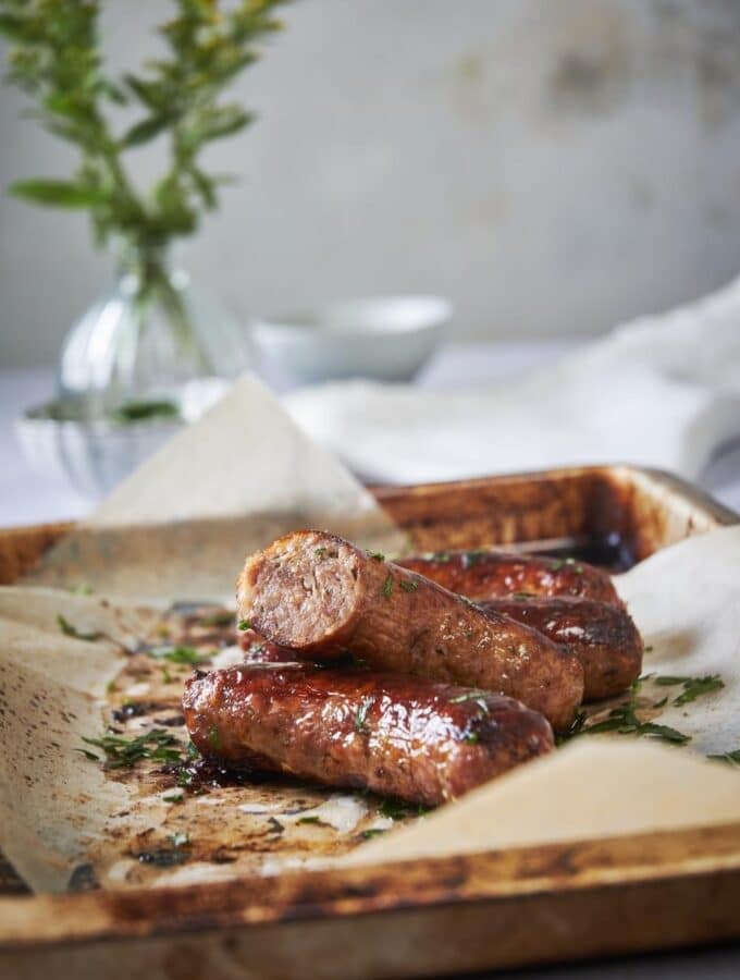 Four brats on a sheet pan lined with parchment paper garnished with parsley. One of the brats is cut in half to show the pink and juicy cross section.